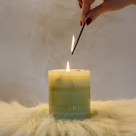 Hand lighting a candle with the brand 'BOUJEE' on a fluffy surface.